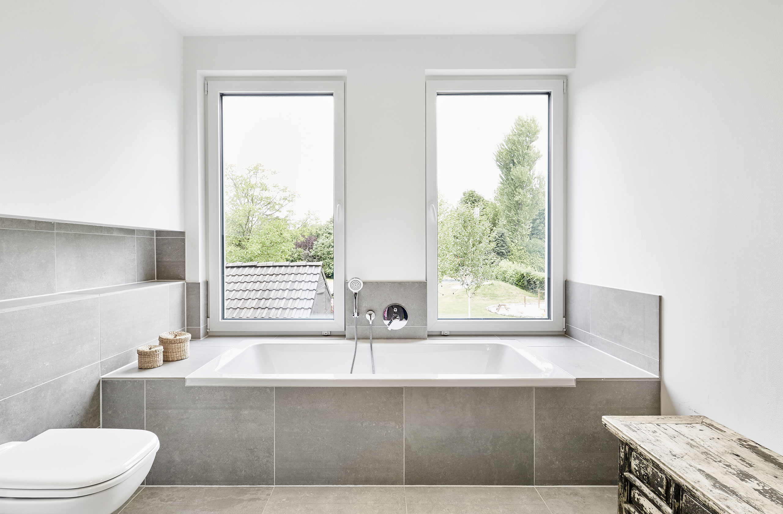 Badezimmer mit Wanne, grauen Fliesen und zwei Fenstern