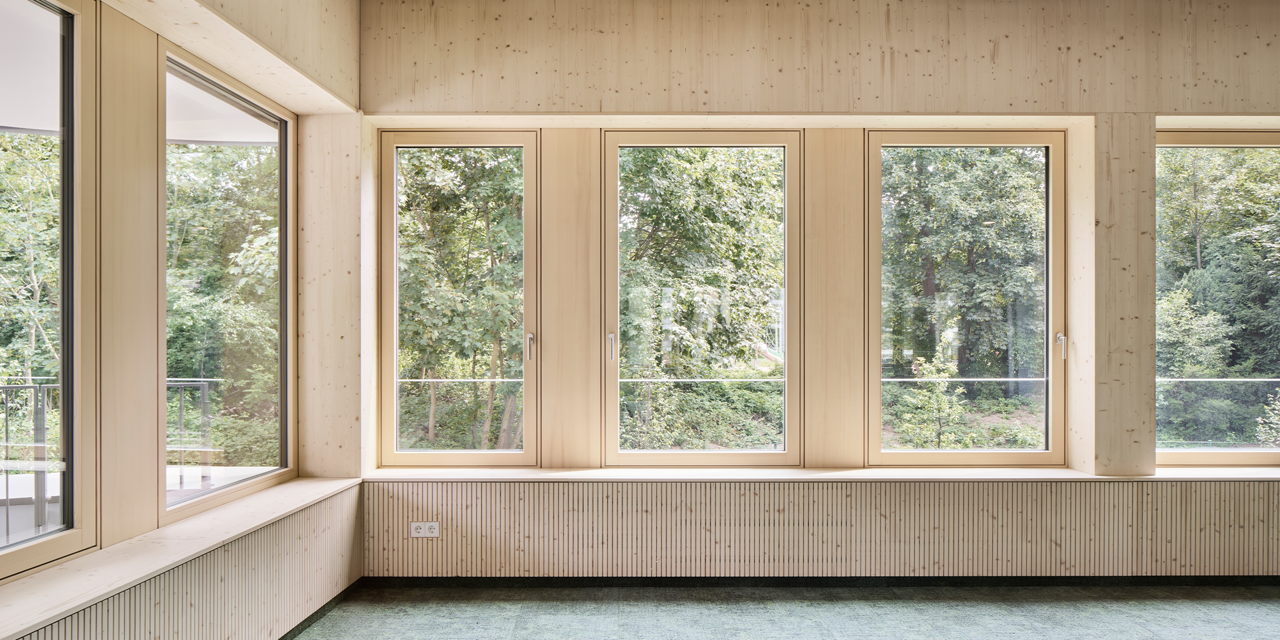 Heller Innenraum mit sichtbaren Holzoberflächen, großen Fenstern und Deckenelementen. Die raumhohen Fenster geben den Blick auf einen dicht bewachsenen Baumbestand frei und sorgen für viel Tageslicht.